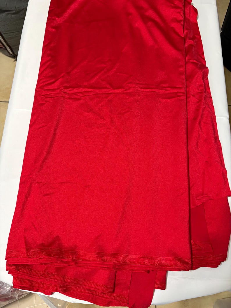 May include: A length of vibrant red fabric with a smooth, satin-like finish. The material is folded in layers, displaying a consistent, solid red color throughout. The edges show a subtle, fine-ribbed texture along the hem of the textile.