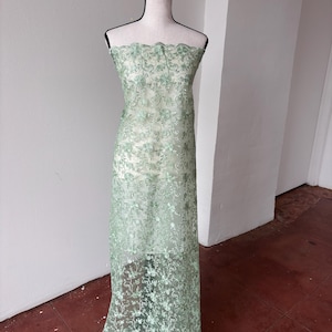 Op de afbeelding: Lichtgroene kanten stof met een bloemmotief, gedrapeerd over een mannequin. De stof heeft een geschulpte rand en is semi-transparant. Het kant is lang, geschikt voor jurken of andere kledingstukken. Afmetingen in centimeters.