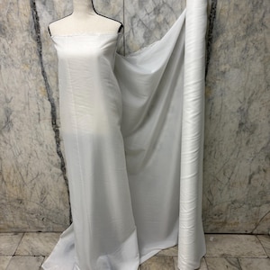 May include: A white fabric roll and draped fabric on a mannequin. The fabric is a solid white color and appears to be a lightweight material. The mannequin is a neutral color and is positioned in front of a marble-patterned wall.