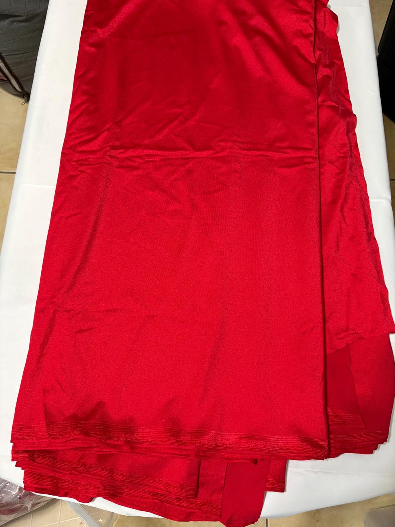 May include: A length of solid red fabric with a smooth, slightly reflective surface texture. The material is folded in layers, showing a consistent vibrant red color throughout the entire piece. The edges appear finished with a subtle horizontal ribbed pattern.