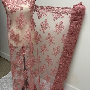 K&ouml;nnte beinhalten: Staubrosa Spitzenstoff mit floraler Stickerei. Der Stoff ist &uuml;ber eine Schaufensterpuppe drapiert und aufgerollt, wodurch die komplizierten Details des Designs zur Geltung kommen. Die Spitze hat einen gewellten Rand und ein zartes, transparentes Aussehen.