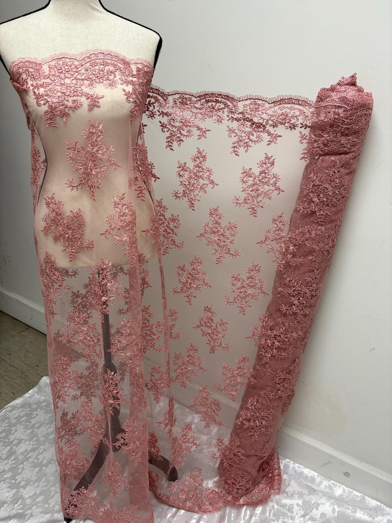 K&ouml;nnte beinhalten: Staubrosa Spitzenstoff mit floraler Stickerei. Der Stoff ist &uuml;ber eine Schaufensterpuppe drapiert und aufgerollt, wodurch die komplizierten Details des Designs zur Geltung kommen. Die Spitze hat einen gewellten Rand und ein zartes, transparentes Aussehen.
