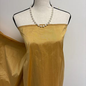 May include: A length of golden yellow fabric with a subtle slubbed texture draped over a mannequin. The fabric has a slight sheen and is paired with a single strand of white round pearls resting around the neck area of the mannequin form.