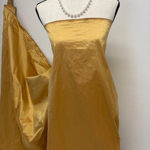 May include: A length of solid gold-colored fabric draped over a mannequin, featuring a smooth, slightly lustrous finish. A single strand of white pearl beads is positioned around the neck area of the mannequin, resting above the fabric.