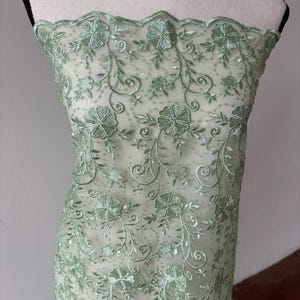 Op de afbeelding: Een doorschijnende, mintgroene stof met geborduurde bloemen- en bladerpatronen. Het delicate ontwerp heeft gedetailleerde steken van bloemen en wijnranken, waardoor een gestructureerd, decoratief oppervlak ontstaat. De stof is over een mannequin gedrapeerd en toont zijn potentieel voor kleding- of knutselprojecten.
