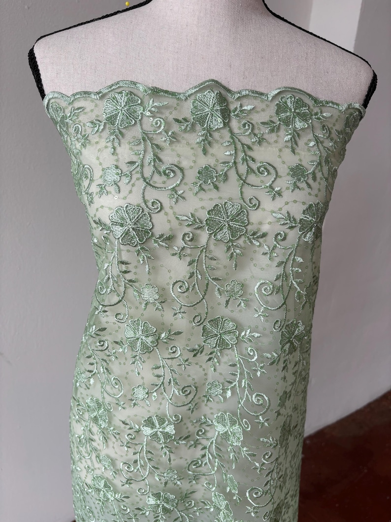 Op de afbeelding: Een doorschijnende, mintgroene stof met geborduurde bloemen- en bladerpatronen. Het delicate ontwerp heeft gedetailleerde steken van bloemen en wijnranken, waardoor een gestructureerd, decoratief oppervlak ontstaat. De stof is over een mannequin gedrapeerd en toont zijn potentieel voor kleding- of knutselprojecten.