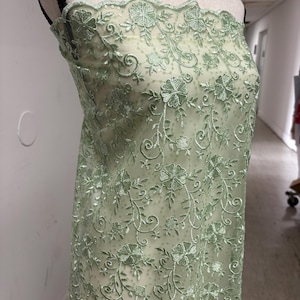 Op de afbeelding: Lichtgroene bloemenkanten stof, getoond op een mannequin. Het kant heeft een geborduurd ontwerp van bloemen en wijnranken. De stof heeft een geschulpte rand en een delicate, transparante uitstraling, geschikt voor kleding of decoratieve projecten.