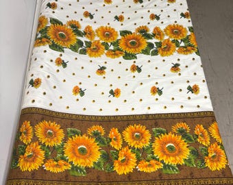 Peut inclure: Un tissu blanc avec un motif répété de tournesols jaunes, oranges et verts. Le tissu a des bordures marron avec des motifs de tournesols et de petits points dorés. Le tissu est enroulé.