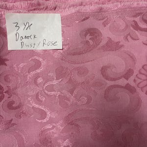 May include: Dusty rose damask fabric with a floral pattern. The fabric is folded and stacked, with a handwritten label that reads "3 Yd Damask Dusty Rose."