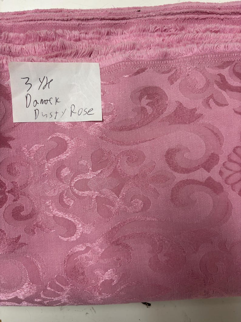May include: Dusty rose damask fabric with a floral pattern. The fabric is folded and stacked, with a handwritten label that reads "3 Yd Damask Dusty Rose."