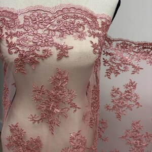 K&ouml;nnte beinhalten: Staubrosa Spitzenstoff mit floraler Stickerei. Die zarte Spitze ist &uuml;ber eine Schaufensterpuppe drapiert und zeigt die komplizierten Blumenmuster und die gewellten Kanten. Der Stoff ist halbtransparent.
