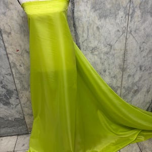 May include: A bolt of bright chartreuse fabric draped over a mannequin. The fabric has a slight sheen and a soft, flowing drape. The top edge of the fabric is unfinished, with a raw, fringed edge. The background is a marble-patterned wall.
