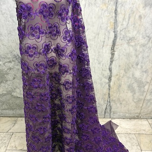 May include: A sheer purple fabric with floral embroidery and sequin accents. The fabric is draped, showcasing the intricate details of the embroidered flowers. The design is a repeating pattern of flowers.
