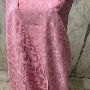 May include: A close-up of pink fabric with a detailed floral pattern. The fabric is folded, revealing the texture and design. The color is a soft, light pink, and the fabric appears to be a luxurious material.