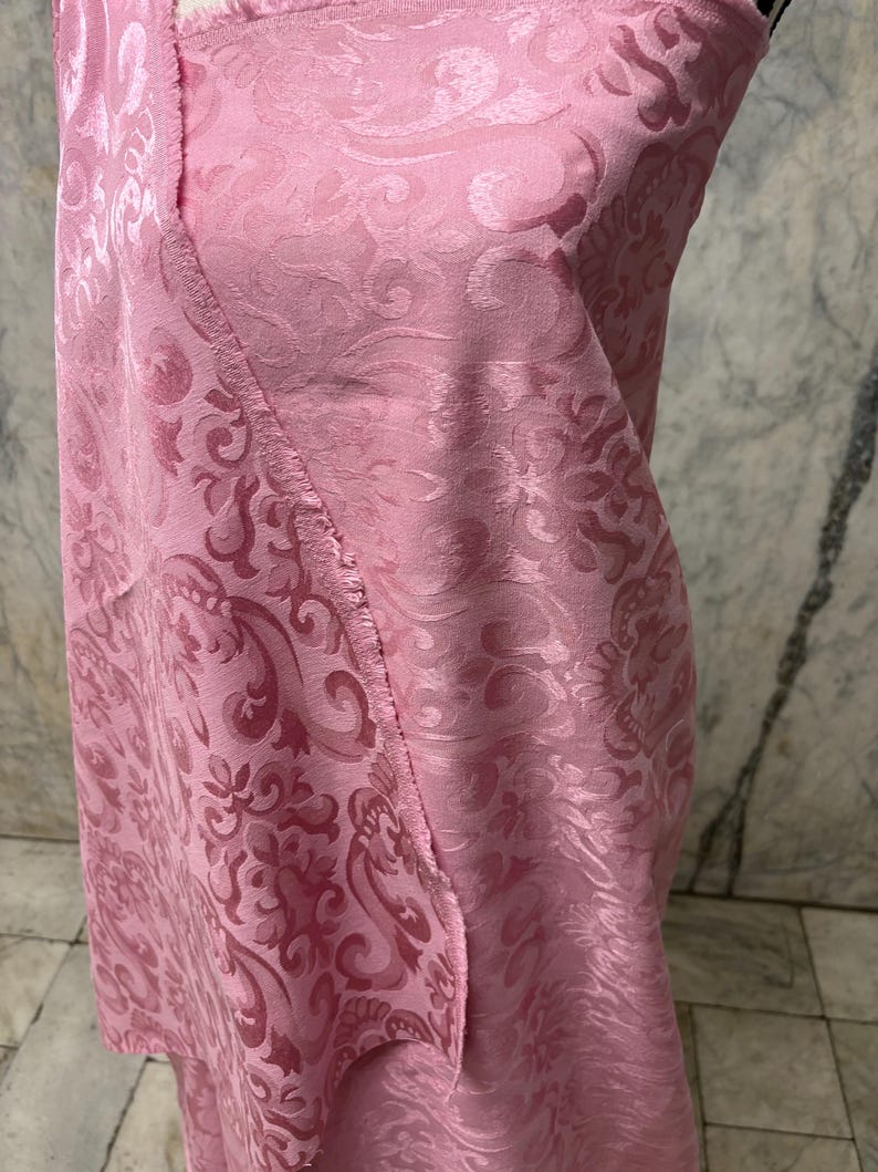 May include: A close-up of pink fabric with a detailed floral pattern. The fabric is folded, revealing the texture and design. The color is a soft, light pink, and the fabric appears to be a luxurious material.