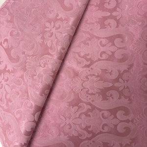 May include: A folded piece of pink fabric with a detailed, raised floral pattern. The fabric has a subtle sheen, and the design includes swirling elements and stylized flowers. The edges of the fabric are visible, showing the texture and weave.