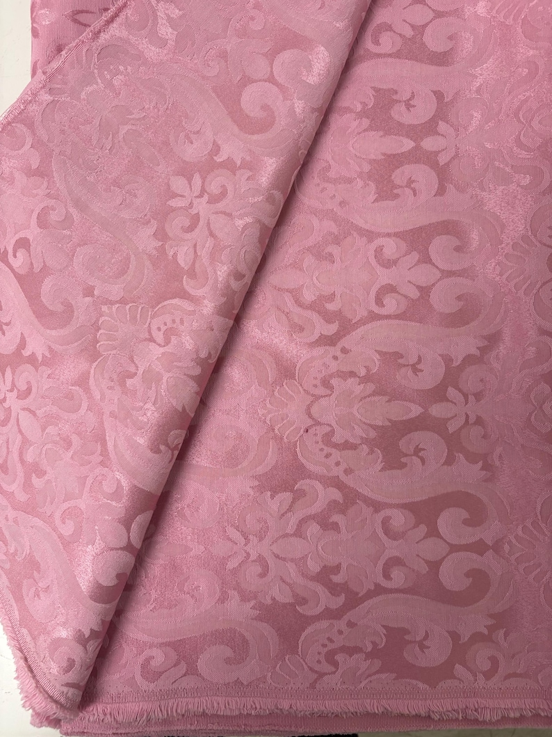 May include: A folded piece of pink fabric with a detailed, raised floral pattern. The fabric has a subtle sheen, and the design includes swirling elements and stylized flowers. The edges of the fabric are visible, showing the texture and weave.