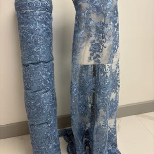 May include: A roll of blue lace fabric and a dress form draped with matching lace. The lace features a floral pattern with scalloped edges. The fabric is a light blue color and appears to be semi-sheer, suitable for dresses or other apparel.