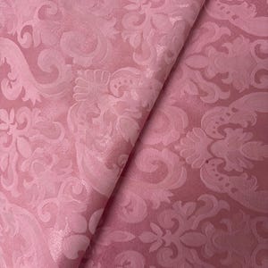 May include: A folded piece of pink fabric with a detailed floral damask pattern. The fabric has a subtle sheen, and the design features swirling leaves and flowers. The color is a soft, muted pink, and the fabric appears to be a medium weight.