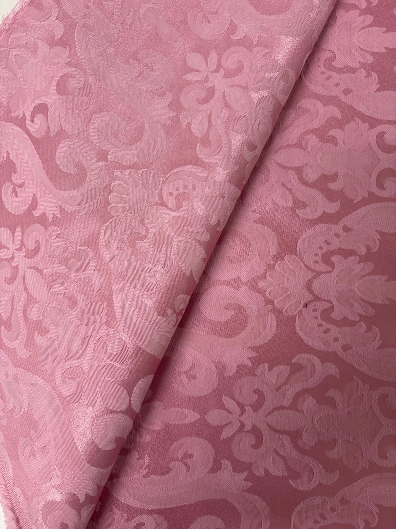 May include: A folded piece of pink fabric with a detailed floral damask pattern. The fabric has a subtle sheen, and the design features swirling leaves and flowers. The color is a soft, muted pink, and the fabric appears to be a medium weight.