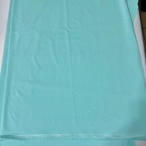 May include: A solid light turquoise fabric with a smooth, matte texture. The material is folded in layers, displaying a consistent, uniform color throughout the surface. The edges of the fabric show a clean, straight finish along the bottom.