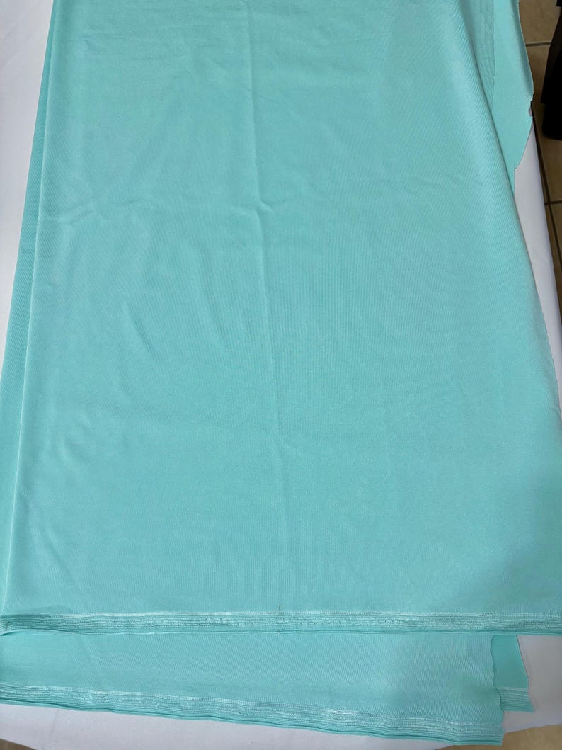 May include: A solid light turquoise fabric with a smooth, matte texture. The material is folded in layers, displaying a consistent, uniform color throughout the surface. The edges of the fabric show a clean, straight finish along the bottom.