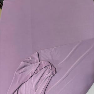 May include: A solid, light purple fabric with a smooth, matte texture. The material is draped to show a folded corner, revealing a consistent, opaque color throughout the surface. The fabric appears lightweight and flexible with soft folds.