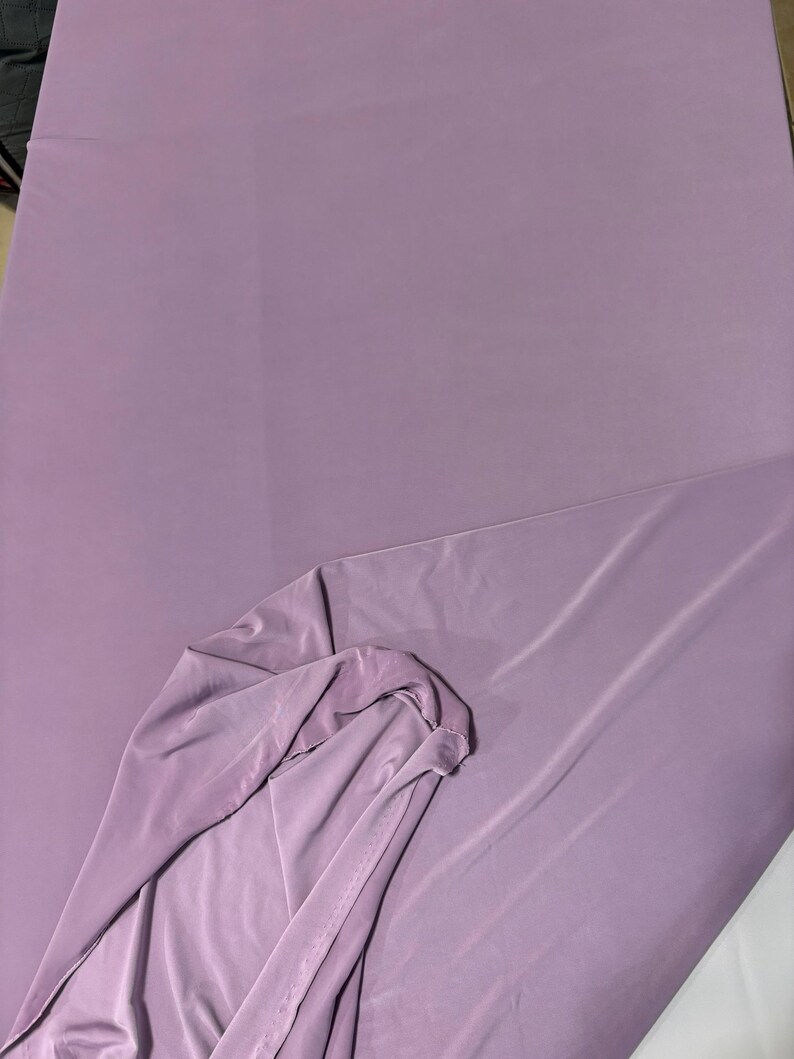 May include: A solid, light purple fabric with a smooth, matte texture. The material is draped to show a folded corner, revealing a consistent, opaque color throughout the surface. The fabric appears lightweight and flexible with soft folds.