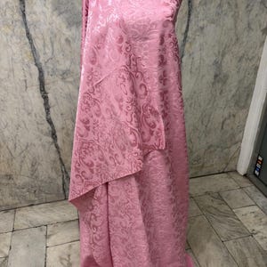 May include: Pink fabric draped over a mannequin. The fabric has a floral pattern and a textured surface. The fabric is a light pink color and appears to be a medium weight material. The fabric is likely used for clothing or home decor.