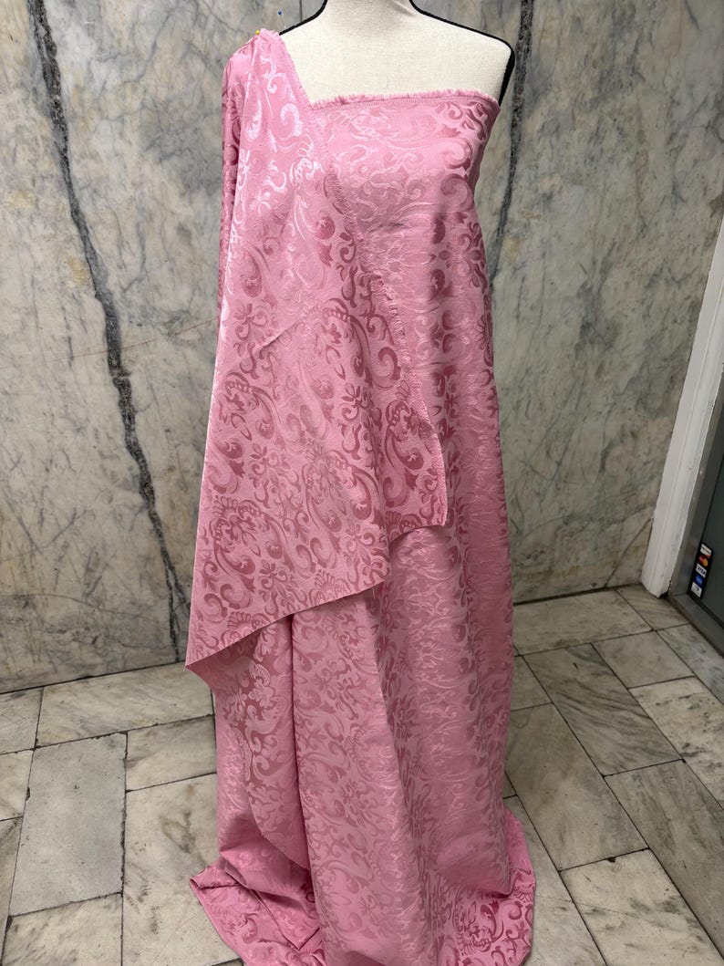 May include: Pink fabric draped over a mannequin. The fabric has a floral pattern and a textured surface. The fabric is a light pink color and appears to be a medium weight material. The fabric is likely used for clothing or home decor.