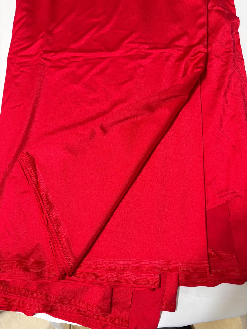 May include: A vibrant red fabric with a smooth, slightly reflective surface. The material is folded over itself, creating soft drapes and shadows that highlight the consistent, solid red color and the fine, woven texture of the textile edges.