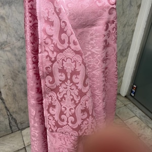 May include: Pink fabric with a damask pattern. The fabric is a light pink color with a repeating floral and scroll design. The fabric is draped over a form, showing the texture and pattern.