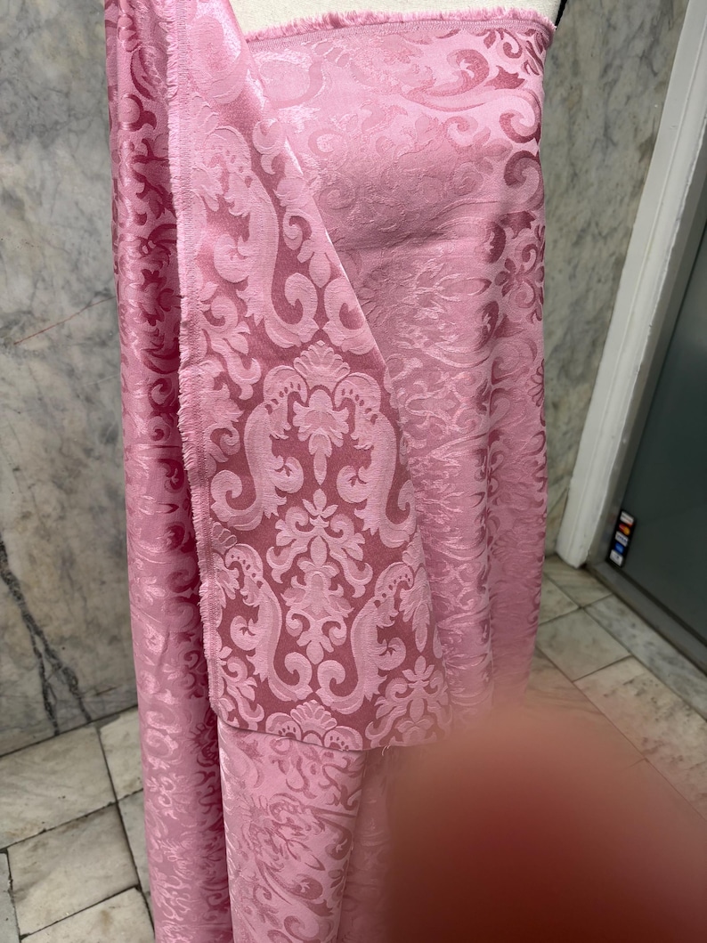 May include: Pink fabric with a damask pattern. The fabric is a light pink color with a repeating floral and scroll design. The fabric is draped over a form, showing the texture and pattern.