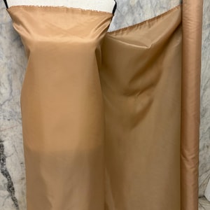May include: Tan-colored fabric draped over a mannequin and rolled up. The fabric has a smooth, shiny texture and a raw edge. The background is a marble-like pattern. This fabric is suitable for apparel and craft projects.