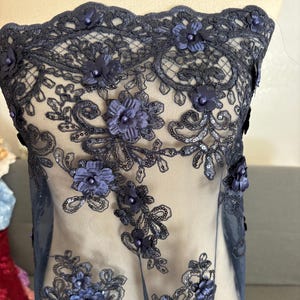 May include: Navy blue lace fabric with 3D floral embellishments. The delicate lace features intricate patterns and is adorned with clusters of small, dark blue flowers and sequins. The fabric is semi-sheer, revealing a neutral background.
