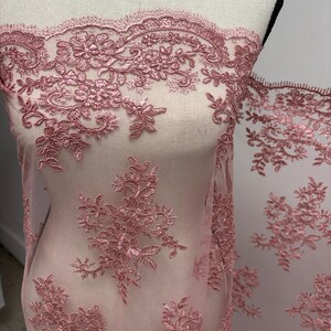 K&ouml;nnte beinhalten: Staubrosa Spitzenstoff mit floraler Stickerei. Die zarte Spitze weist detaillierte Blumen- und Bl&auml;ttermuster auf. Der Stoff ist &uuml;ber eine Schaufensterpuppe drapiert und zeigt das komplizierte Design und die Textur. Ideal f&uuml;r Brautmode oder besondere Anl&auml;sse.