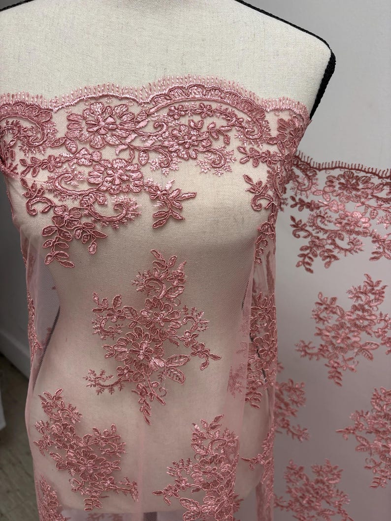 K&ouml;nnte beinhalten: Staubrosa Spitzenstoff mit floraler Stickerei. Die zarte Spitze weist detaillierte Blumen- und Bl&auml;ttermuster auf. Der Stoff ist &uuml;ber eine Schaufensterpuppe drapiert und zeigt das komplizierte Design und die Textur. Ideal f&uuml;r Brautmode oder besondere Anl&auml;sse.