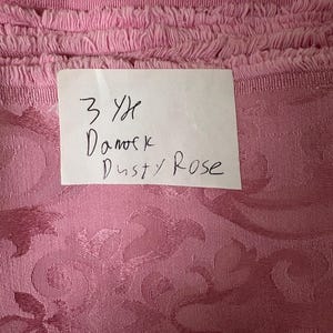 May include: Close-up of a roll of dusty rose fabric with a floral damask pattern. The top edge has a fringed trim. A handwritten note reads "3 Yd Damask Dusty Rose."