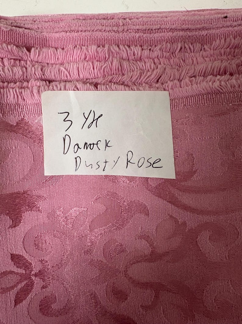 May include: Close-up of a roll of dusty rose fabric with a floral damask pattern. The top edge has a fringed trim. A handwritten note reads "3 Yd Damask Dusty Rose."