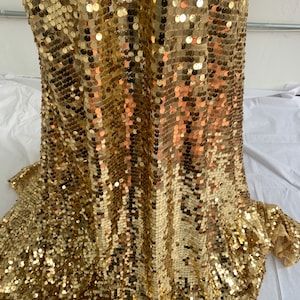 Może przedstawiać: Tkanina z cekinów w kolorze złotym z subtelnym połyskiem. Tkanina wykonana jest z małych, okrągłych cekinów, które są zszyte razem w ciasnym wzorze. Cękiny są jasnozłote i mają delikatny, tęczowy połysk.