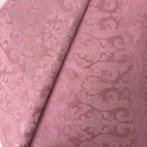 May include: A folded piece of pink fabric with a detailed floral and scroll pattern. The fabric has a slight sheen and a soft texture, with the pattern slightly raised. The edges of the fabric are frayed.