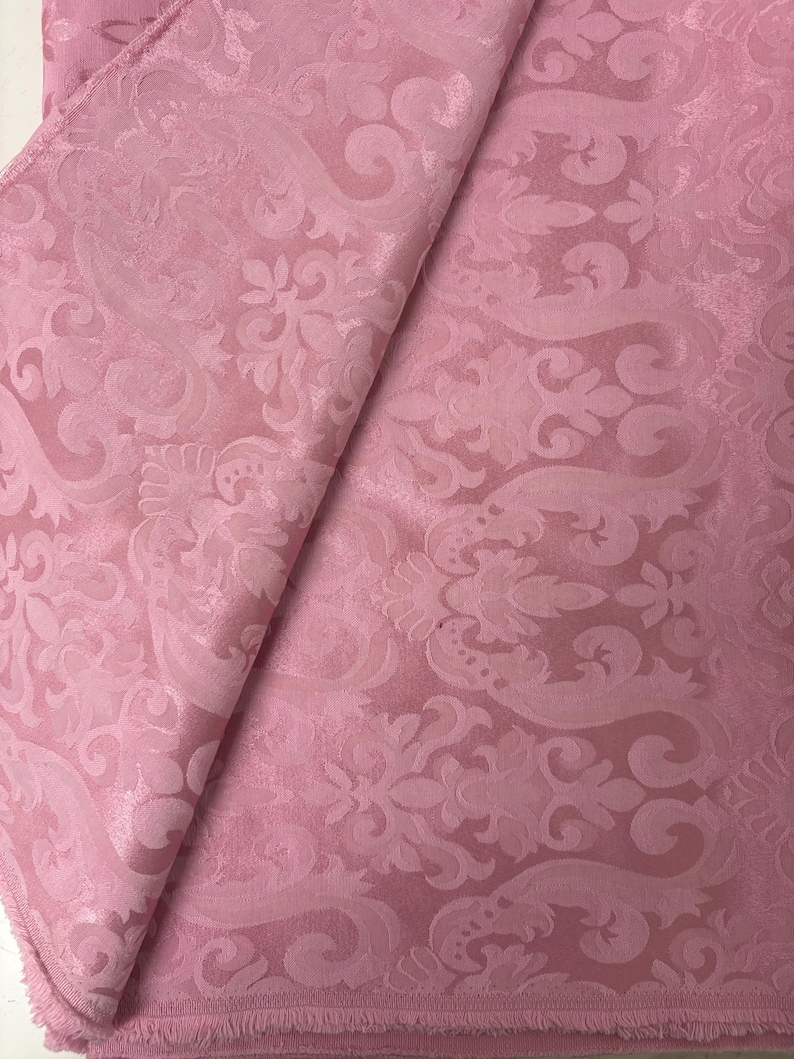 May include: A folded piece of pink fabric with a detailed floral and scroll pattern. The fabric has a slight sheen and a soft texture, with the pattern slightly raised. The edges of the fabric are frayed.