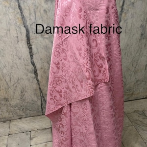 May include: Pink damask fabric draped over a mannequin. The fabric has a repeating floral pattern and the words "Damask fabric" are printed on the fabric. The fabric is a light pink color.