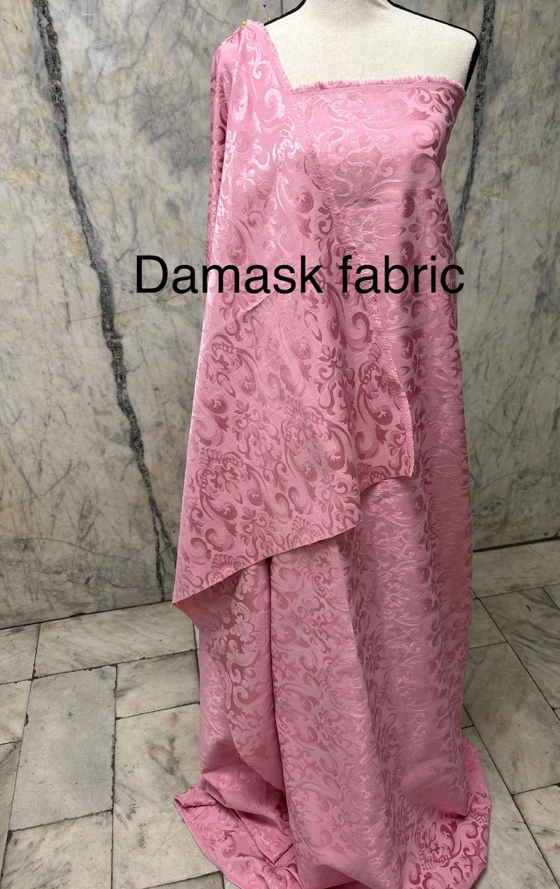 May include: Pink damask fabric draped over a mannequin. The fabric has a repeating floral pattern and the words "Damask fabric" are printed on the fabric. The fabric is a light pink color.