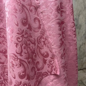 May include: Close-up of a pink fabric with a detailed damask pattern. The fabric has a soft sheen and a textured surface. The design features swirling floral and scroll motifs, creating a luxurious look. The edge of the fabric is visible.