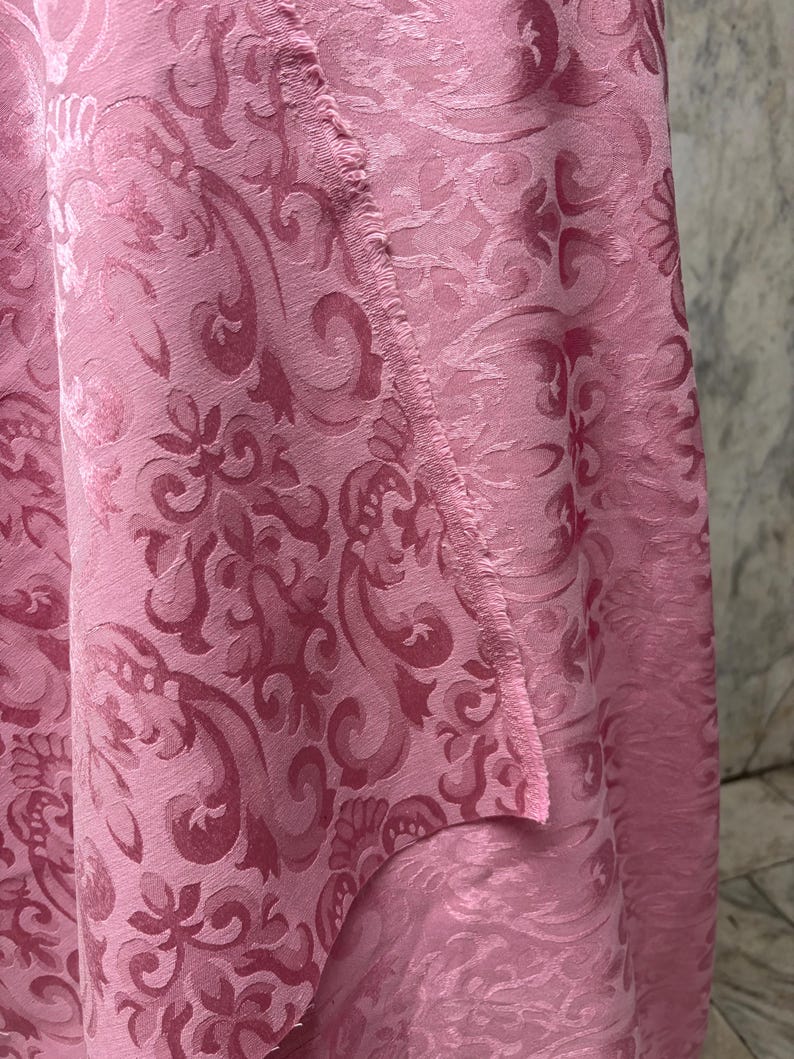 May include: Close-up of a pink fabric with a detailed damask pattern. The fabric has a soft sheen and a textured surface. The design features swirling floral and scroll motifs, creating a luxurious look. The edge of the fabric is visible.