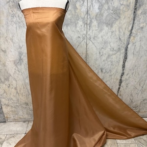 May include: A mannequin draped in a flowing, golden-brown fabric. The fabric is smooth and appears to be a lightweight material, possibly silk or a similar textile. The fabric is draped to create a dress-like form.