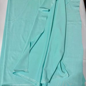 May include: A length of solid light mint green fabric with a smooth, matte texture. The material is draped to show its soft, flexible quality, with visible finished edges along the bottom and a clean, uniform color throughout the entire piece.