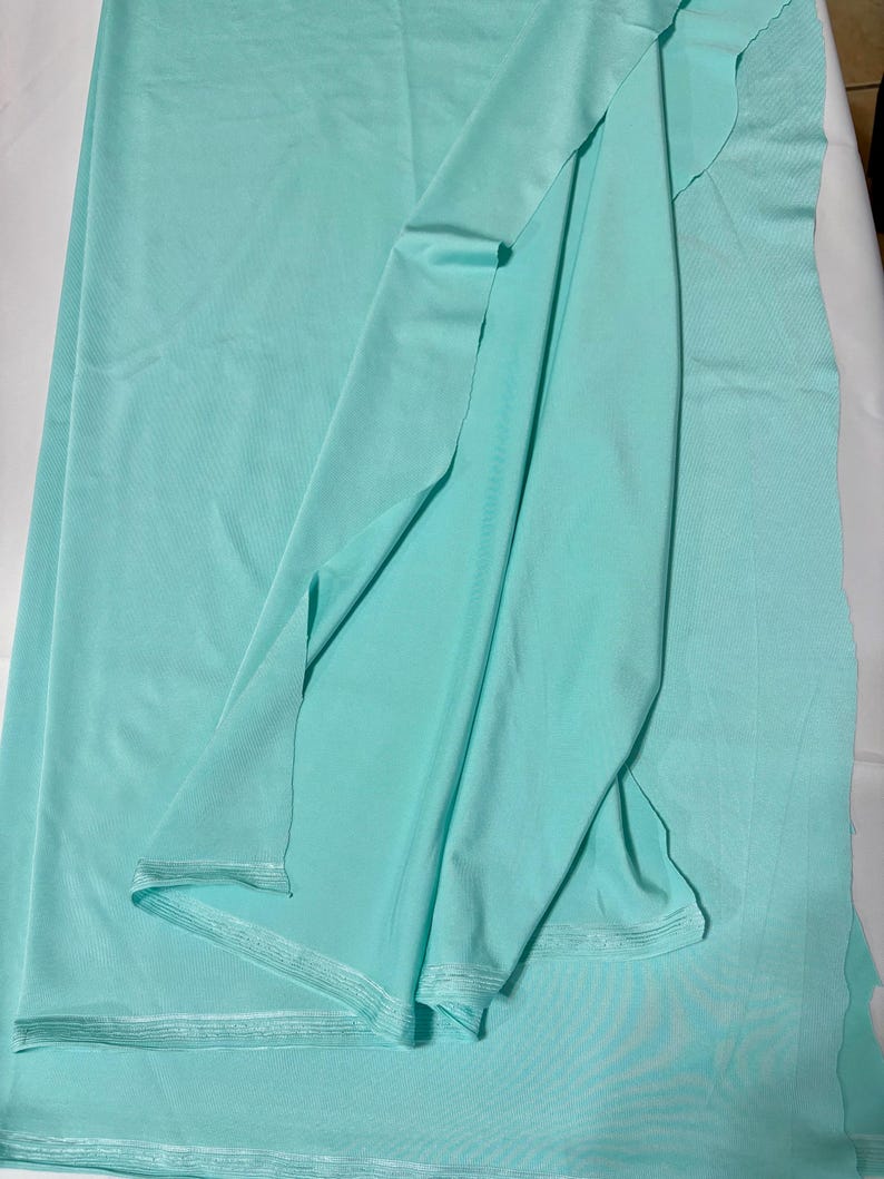 May include: A length of solid light mint green fabric with a smooth, matte texture. The material is draped to show its soft, flexible quality, with visible finished edges along the bottom and a clean, uniform color throughout the entire piece.