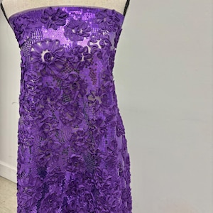 May include: A bolt of purple fabric with a floral design. The fabric is covered in three-dimensional flower appliques and sequins. The fabric is draped over a mannequin.